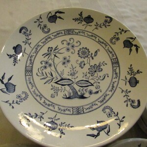 May include: A white ceramic dinner plate with a blue floral design. The design features a central tree with leaves and flowers, surrounded by a border of smaller floral motifs.