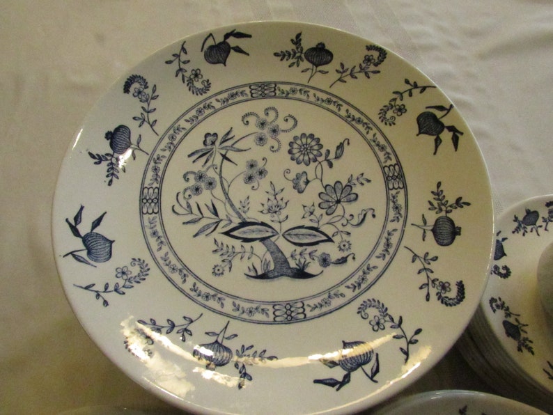 May include: A white ceramic dinner plate with a blue floral design. The design features a central tree with leaves and flowers, surrounded by a border of smaller floral motifs.