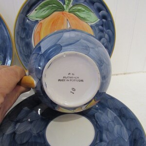 May include: A blue and white ceramic teacup and saucer set with a peach design. The saucer has a scalloped edge and the cup has a handle. The bottom of the cup says "P.B. Alcoba&ccedil;a Made in Portugal 10".