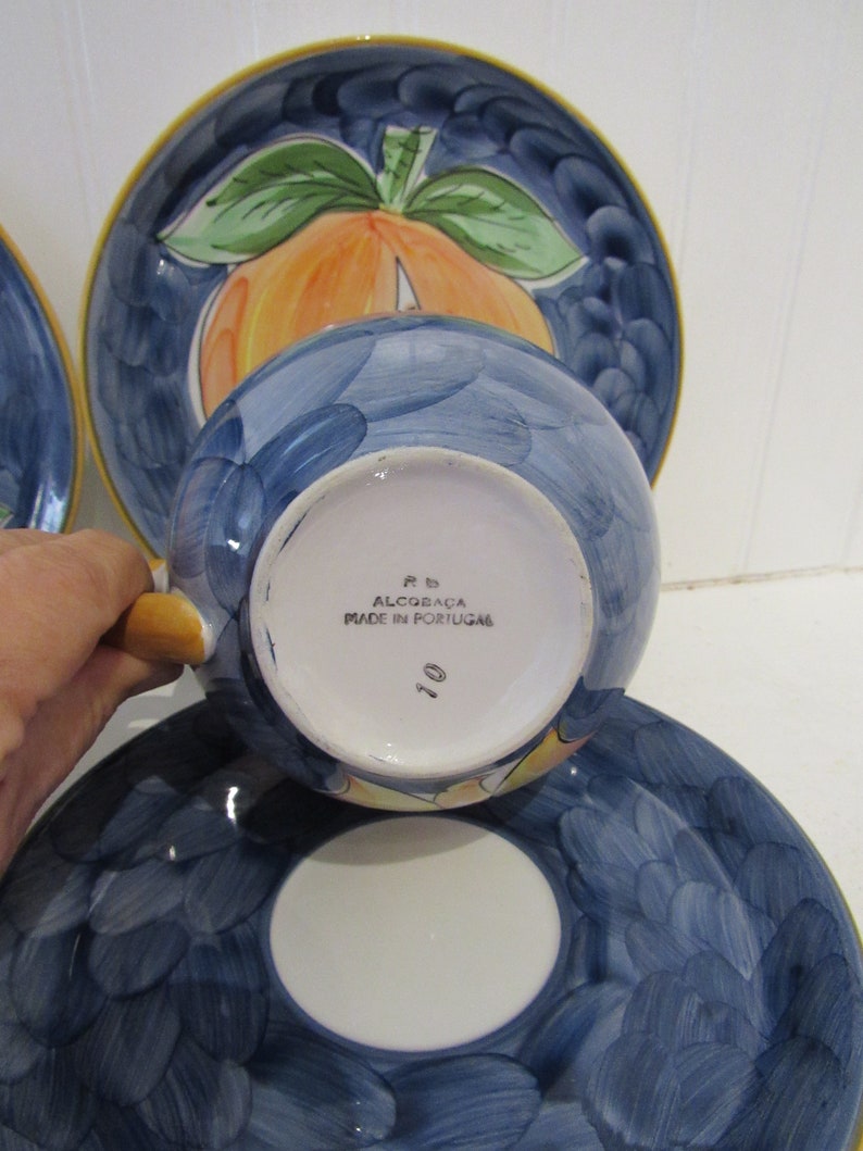 May include: A blue and white ceramic teacup and saucer set with a peach design. The saucer has a scalloped edge and the cup has a handle. The bottom of the cup says "P.B. Alcoba&ccedil;a Made in Portugal 10".