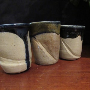 Puede incluir: Tres pequeñas tazas de cerámica con un esmalte blanco y un borde negro. Las tazas tienen un aspecto texturizado, hecho a mano.