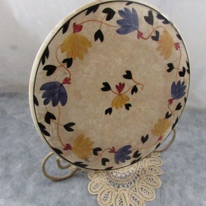 May include: A decorative plate with a floral design. The plate features yellow and blue flowers with black leaves and stems, arranged around the rim. The plate is displayed on a metal stand and rests on a lace doily.