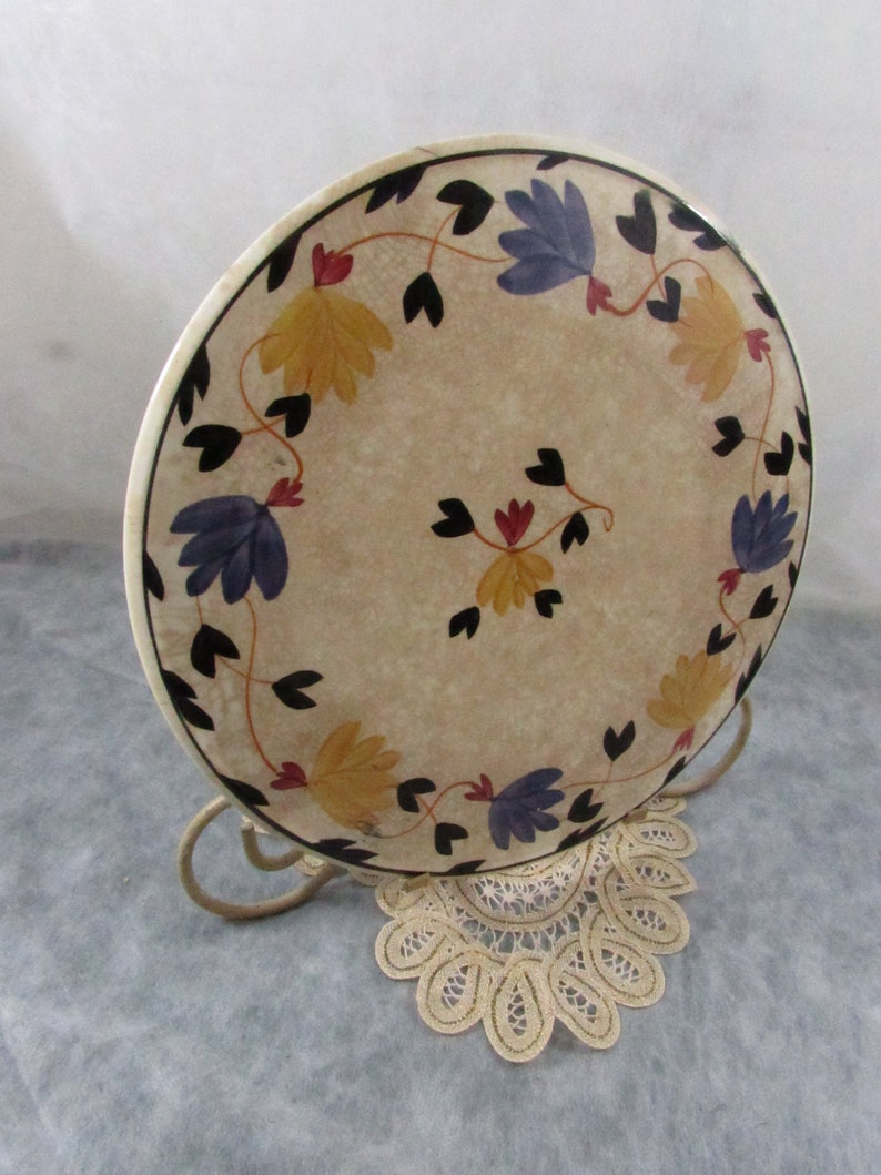 May include: A decorative plate with a floral design. The plate features yellow and blue flowers with black leaves and stems, arranged around the rim. The plate is displayed on a metal stand and rests on a lace doily.