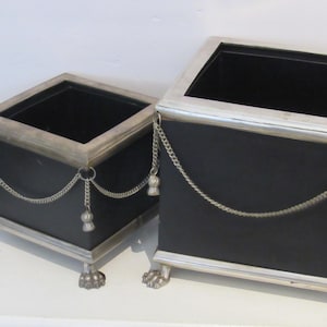 May include: Two black square planters with silver trim and decorative chains. The planters have silver paw feet and a silver trim around the top. The planters are empty and ready for plants.