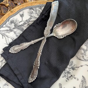 May include: A silver spoon and butter knife rest on a black napkin atop a decorative plate with a floral design. The plate has a gold rim. The silverware has ornate detailing.