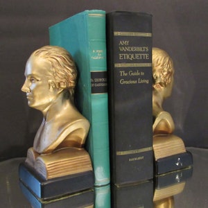 May include: A pair of gold bookends featuring busts of men, holding open books. The bookends are holding a teal book titled "Fr. Leopold of Castelno" and a black book titled "Amy Vanderbilt's Etiquette: The Guide to Gracious Living".