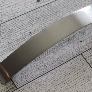 May include: A curved, brushed metal cabinet pull with a rectangular profile. The hardware has a satin finish and is set against a light-colored, wood-grain surface.
