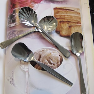 May include: Four silver teaspoons, one with a decorative shell design, are arranged on a white surface with a cup of ice cream and a plate of toast. The spoons are all different sizes and shapes. The ice cream is a light brown color and is topped with a dollop of whipped cream. The toast is a light golden brown color and is cut into small squares.