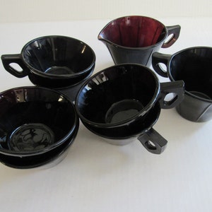 May include: Set of six black glass teacups and a matching creamer. The cups have a fluted design and are stacked in pairs. The creamer has a deep burgundy color.