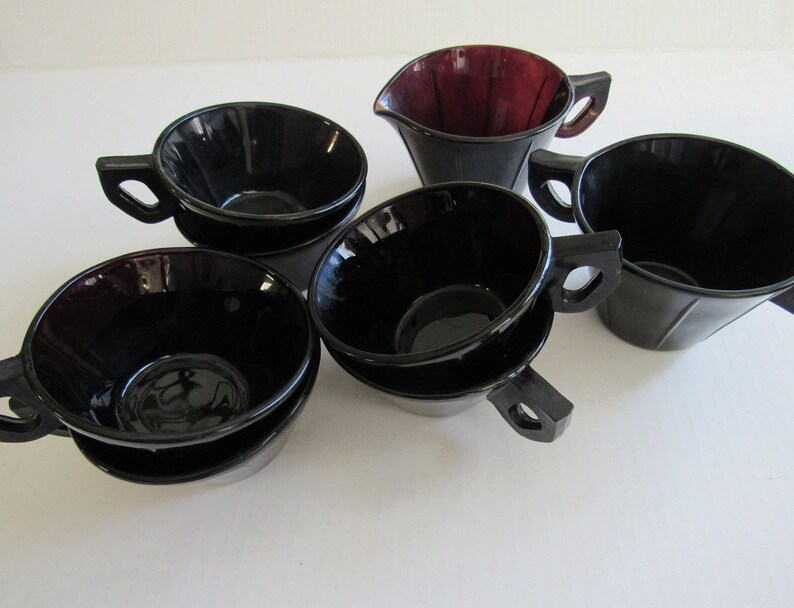 May include: Set of six black glass teacups and a matching creamer. The cups have a fluted design and are stacked in pairs. The creamer has a deep burgundy color.