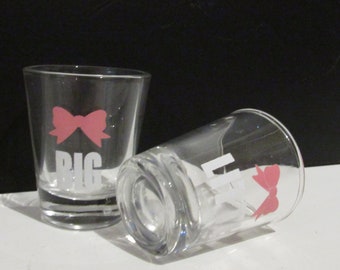 May include: Two clear glass shot glasses with pink bows and white lettering. The first glass says "Big" and the second glass says "Little".