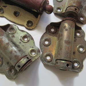 May include: A set of four weathered metal spring hinges with a scalloped edge mounting plate. Each hinge features a cylindrical barrel mechanism and multiple circular screw holes, showing signs of oxidation, rust, and remnants of old paint.