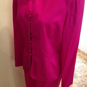 Op de afbeelding: Een fel fuchsia tweedelig pak. De jas heeft een reverskraag en bloemvormige knoopsluitingen. De rok is in een bijpassende effen kleur. Het pak wordt op een mannequin gepresentteerd.