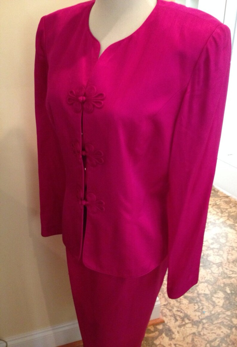 Op de afbeelding: Een fel fuchsia tweedelig pak. De jas heeft een reverskraag en bloemvormige knoopsluitingen. De rok is in een bijpassende effen kleur. Het pak wordt op een mannequin gepresentteerd.