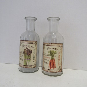 May include: Two clear glass bottles with ribbed sides and decorative labels. The labels feature illustrations of asparagus and carrots with text that reads "Asparagus" and "Carotte".