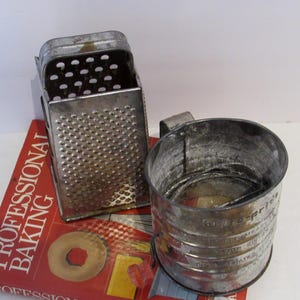 May include: Two vintage metal kitchen tools: a grater and a flour sifter. The grater is rectangular with a perforated surface. The sifter is a cylindrical cup with a handle and a mesh screen. Both tools are placed on a red book titled "PROFESSIONAL BAKING."