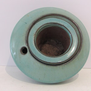 May include: A turquoise ceramic ashtray with a circular design. The ashtray has a central well and a smaller, concentric ring. A small hole is visible on the side. The rim is a darker brown color.