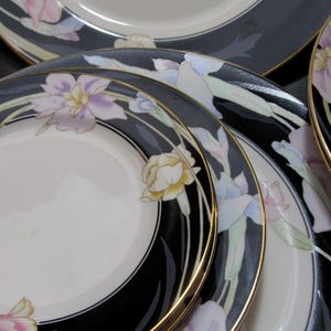 May include: A collection of white ceramic plates with a black border and gold trim. The plates are decorated with floral designs in shades of pink, purple, and yellow. The plates are arranged in a layered composition.