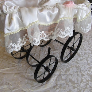 May include: A vintage-style baby carriage with a white satin canopy and delicate lace trim. The carriage features a wicker basket and black metal wheels. Pink rose accents adorn the canopy, adding a touch of elegance to this classic design.