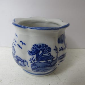 May include: A small, white ceramic pot with a blue, scenic design. The pot features a scalloped rim and a blue border. The blue design depicts trees, buildings, and birds. The pot is likely used for decorative purposes.
