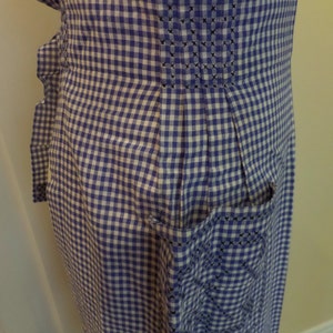 May include: A blue and white gingham apron with a pocket and a pleated back. The apron has a white trim around the edges and a black embroidered design on the pocket.