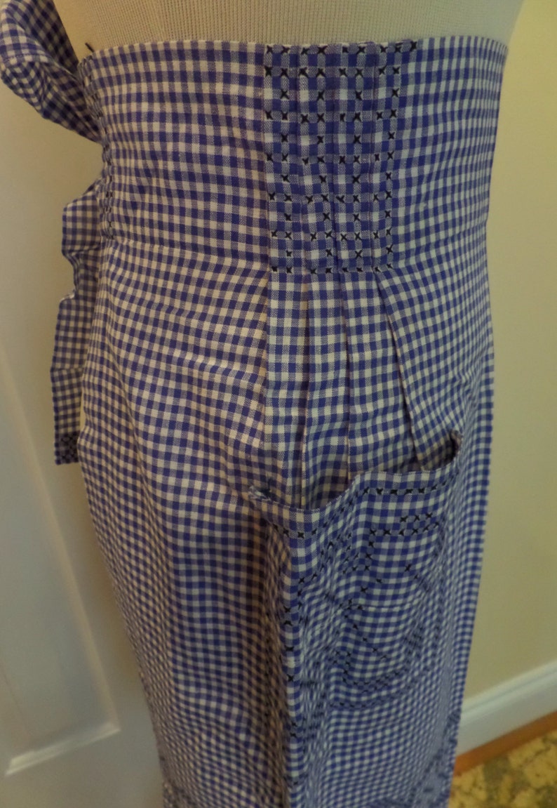 May include: A blue and white gingham apron with a pocket and a pleated back. The apron has a white trim around the edges and a black embroidered design on the pocket.