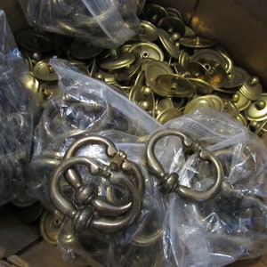 Puede incluir: Una caja llena de una gran cantidad de herrajes para armarios de metal dorado. Los herrajes incluyen pomos redondos y tiradores decorativos.