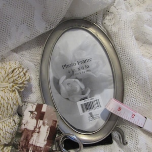 May include: An oval silver photo frame, 4 in. x 6 in., with a black backing, stands on a white lace surface. A vintage photograph and a measuring tape are next to the frame. The frame has a label with the text "Photo Frame".