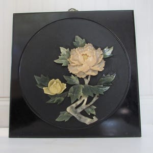 Puede incluir: Una placa de pared negra enmarcada con un diseño floral de jade tallado. Las flores son blancas y amarillas con hojas verdes.