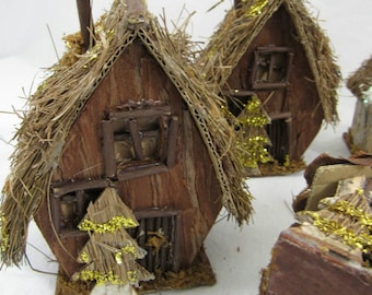 Könnte beinhalten: Drei kleine Holzhäuser mit Strohdächern und goldenen Glitzerakzenten. Jedes Haus hat ein kleines Fenster und eine Tür. Die Häuser hängen an einer Schnur und sind perfekt für Weihnachtsdekorationen.