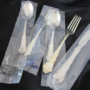 May include: Four pieces of silverware in individual plastic bags. The silverware includes a fork, a knife, and two spoons. The silverware is silver in color and has an ornate design.