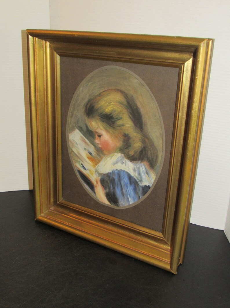 May include: A framed painting of a young girl with blonde hair reading a book. The painting is in an oval shape and is set against a brown background. The frame is gold.