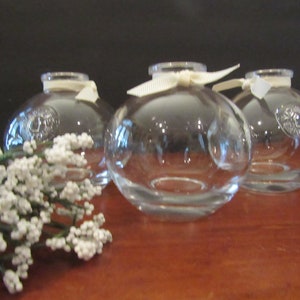 May include: Three clear glass bud vases with white satin ribbon bows. The vases are round and have a slight curve at the bottom.