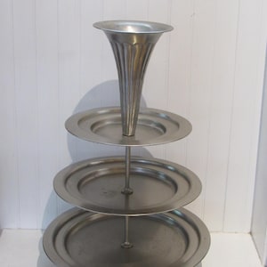 May include: A tiered, silver-toned serving stand with three circular plates and a central, fluted cone. The stand is designed to hold and display items. The base is a larger, circular plate.
