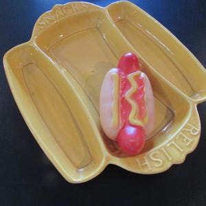 Puede incluir: Una fuente de cerámica amarilla dividida en tres secciones, con las palabras "SNACKS" y "RELISH" en relieve. Un juguete de perrito caliente con detalles rojos, rosas y amarillos se encuentra en el centro.