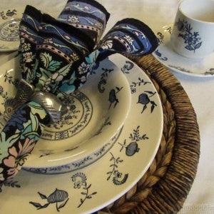 May include: A white ceramic dinner plate with a blue floral pattern, a smaller white ceramic bowl with a blue floral pattern, and a blue and black floral patterned napkin with a silver napkin ring. The plates are on a brown woven placemat.