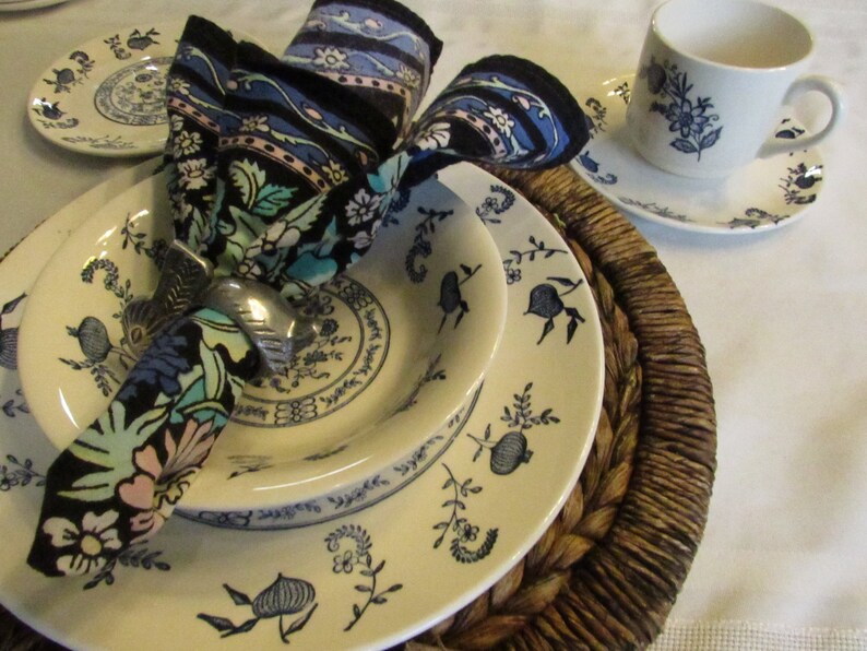 May include: A white ceramic dinner plate with a blue floral pattern, a smaller white ceramic bowl with a blue floral pattern, and a blue and black floral patterned napkin with a silver napkin ring. The plates are on a brown woven placemat.