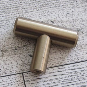 May include: A brass-coloured cabinet pull in a T-shape. The horizontal bar is 7.6 cm long, and the vertical bar is 5.1 cm long. The hardware is set against a light wood grain surface.