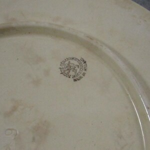 May include: Off-white ceramic plate with a slightly raised rim. A circular emblem with text and a horse-riding figure is visible. The plate has a faded appearance and a number 6 is visible near the bottom.
