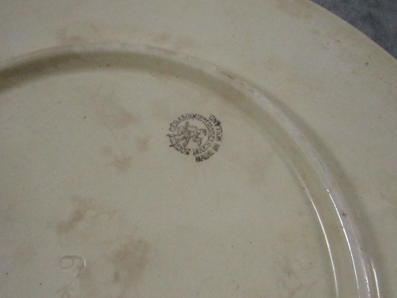 May include: Off-white ceramic plate with a slightly raised rim. A circular emblem with text and a horse-riding figure is visible. The plate has a faded appearance and a number 6 is visible near the bottom.