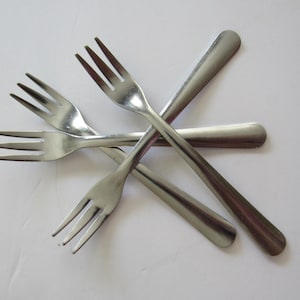 May include: A set of four silver-colored stainless steel forks arranged in a crisscross pattern. Each fork features three tines and a smooth, rounded handle. The metal has a polished, reflective finish, and the forks are positioned overlapping.