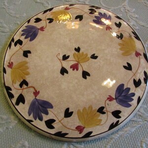 May include: Round ceramic plate with a beige speckled surface, adorned with a floral design. The flowers are in shades of yellow and purple, with black leaves and red accents. The plate has a black rim.