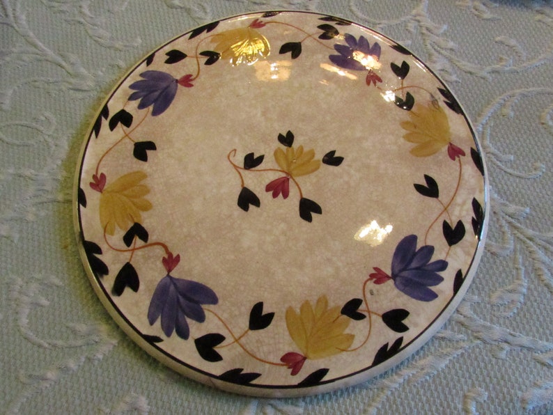 May include: Round ceramic plate with a beige speckled surface, adorned with a floral design. The flowers are in shades of yellow and purple, with black leaves and red accents. The plate has a black rim.