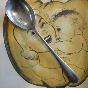 May include: A silver teaspoon with a rounded bowl and a long, slender handle. The spoon is lying on top of a yellow and brown illustration of a mother and child.