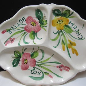 Puede incluir: Una fuente de cerámica blanca con tres secciones, cada una decorada con diseños florales en rosa, amarillo y verde. La fuente está etiquetada con las palabras "Mantequilla", "Queso" y "Lox", con "Bagels" a los lados.
