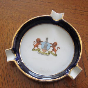 May include: Round ceramic ashtray with a white center featuring a coat of arms design, a navy blue border, and gold trim. The ashtray has three white rests and is set on a wooden surface.