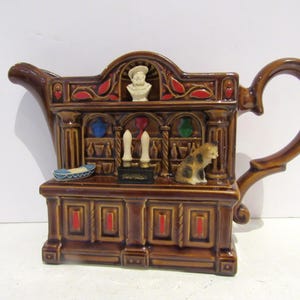 May include: Brown ceramic teapot shaped like a cabinet with a bust, candles, and a small cat figurine. The teapot has a curved handle and spout, with red and blue accents. A small blue and white bowl is on the lower shelf.