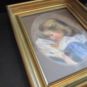May include: A framed painting of a young girl reading a book. The painting is in a gold frame and the girl is wearing a blue dress with a white collar.