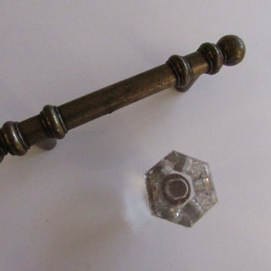 May include: A brass cabinet pull with a rounded knob on each end. The pull has a decorative design of rings around the center. A clear glass knob with a hexagonal shape is also shown.