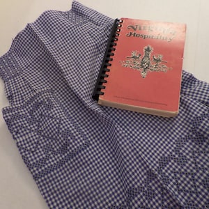 May include: A blue and white gingham fabric apron with a book titled "Virginia Hospitality" on top. The book has a red cover with a pineapple design.